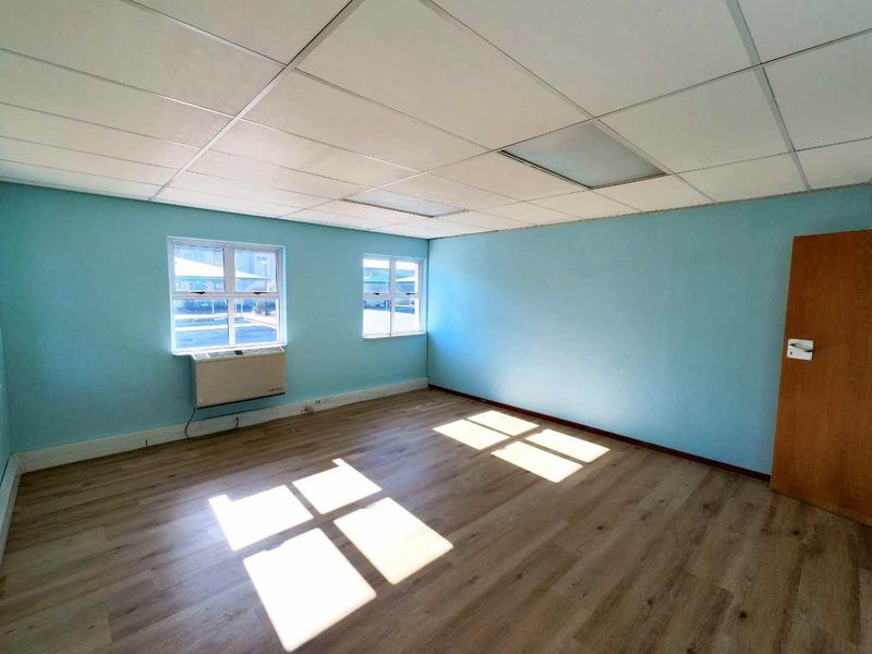 A bright, empty office space featuring laminate flooring, a suspended ceiling, and a built-in window air conditioning unit.