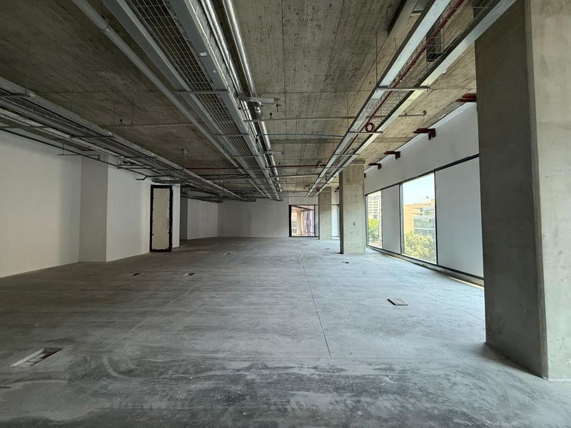 A spacious, open-plan office shell featuring exposed concrete ceilings, industrial ducting, and large windows providing ample natural light.