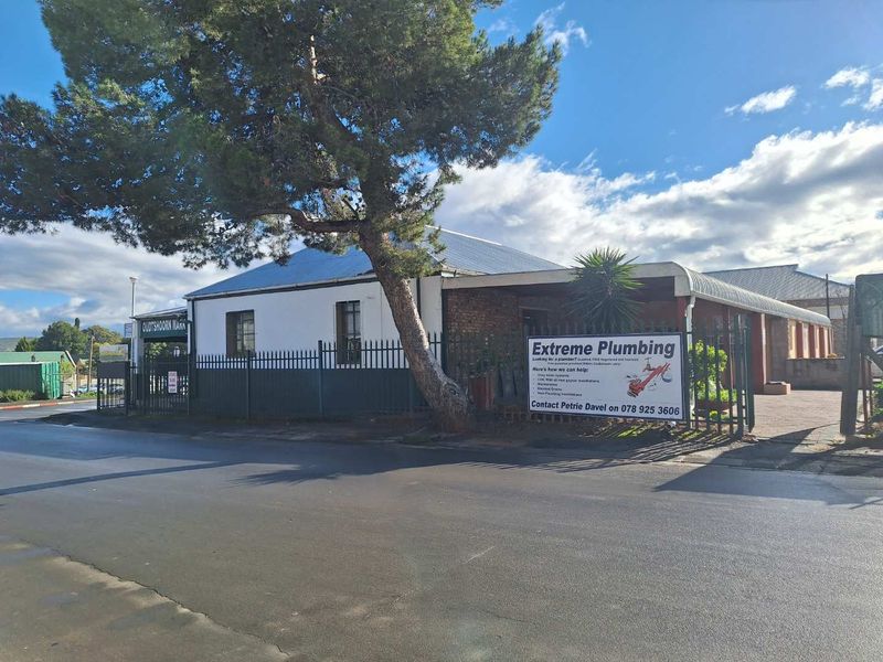 Street-facing view of an industrial property featuring a paved entrance, security fencing, and a corrugated metal roof structure.