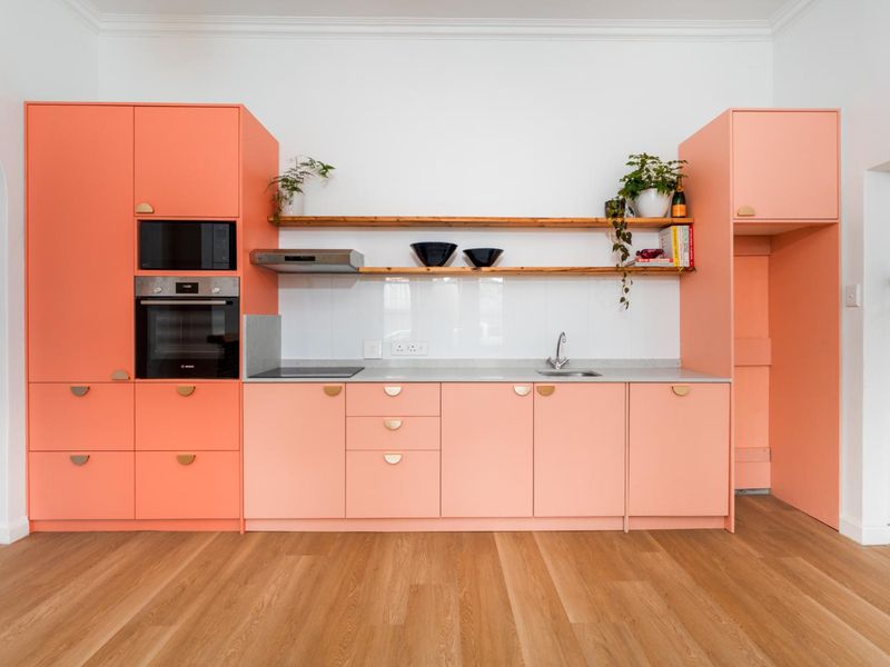 Modern kitchen featuring vibrant coral cabinetry, integrated appliances, timber shelving, and sleek countertops, set against a clean white backdrop with hardwood flooring.