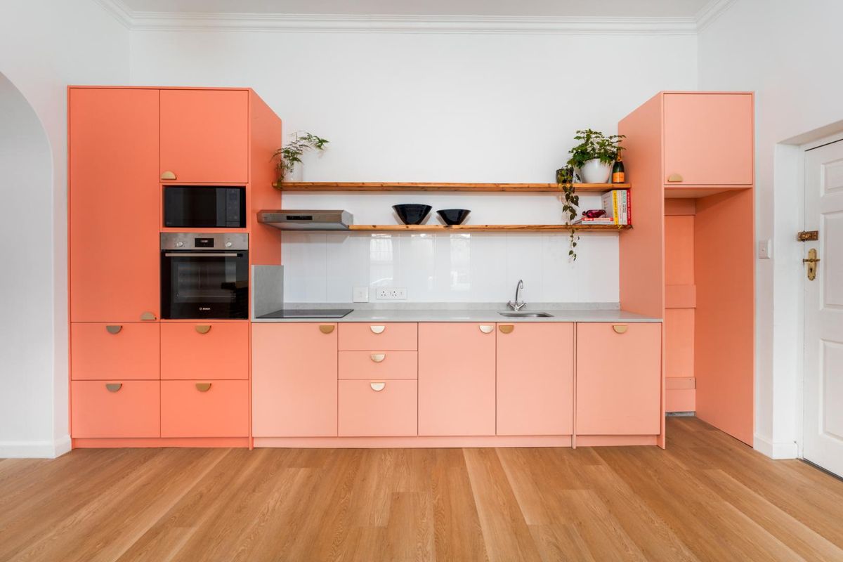Modern kitchen featuring vibrant coral cabinetry, integrated appliances, timber shelving, and sleek countertops, set against a clean white backdrop with hardwood flooring.