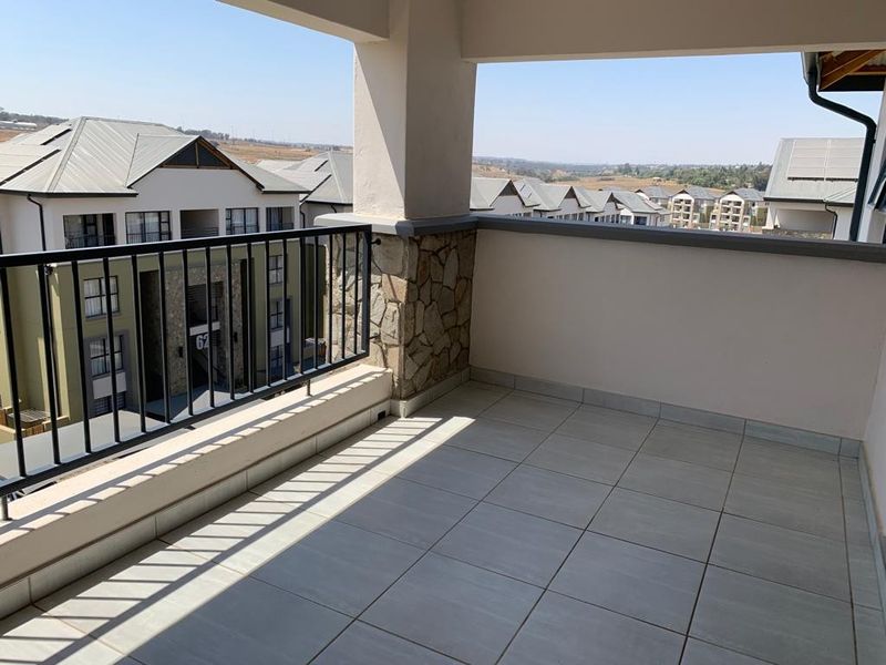 A spacious covered balcony featuring tiled flooring, a metal balustrade, and stone cladding detail, offering an open view of the surrounding residential complex.