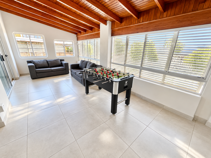 Bright living area featuring exposed timber beam ceilings, large windows with louvre shutters, and neutral tiled flooring, offering a spacious and airy atmosphere.