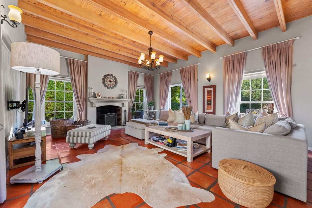 Spacious living room featuring a high exposed timber beam ceiling, traditional masonry fireplace, and classic terracotta floor tiles with large windows.