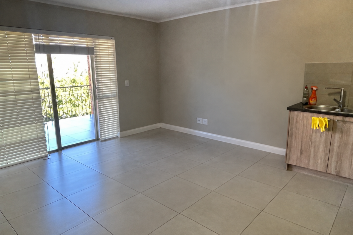 An open-plan living area featuring neutral tiled flooring, a kitchenette with built-in cabinetry, and a sliding glass door leading to a balcony.