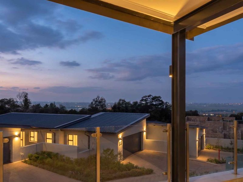 A modern covered balcony featuring a glass balustrade and recessed lighting, offering an elevated view of the surrounding cluster property and landscape at dusk.