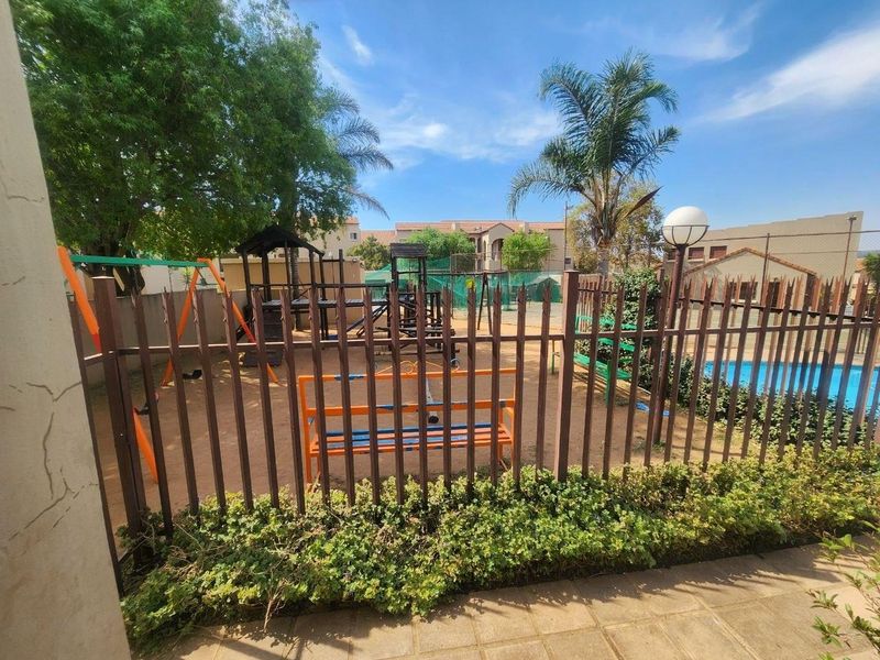 Communal outdoor area featuring a swimming pool, children's playground equipment, and secure metal perimeter fencing within a residential apartment complex.