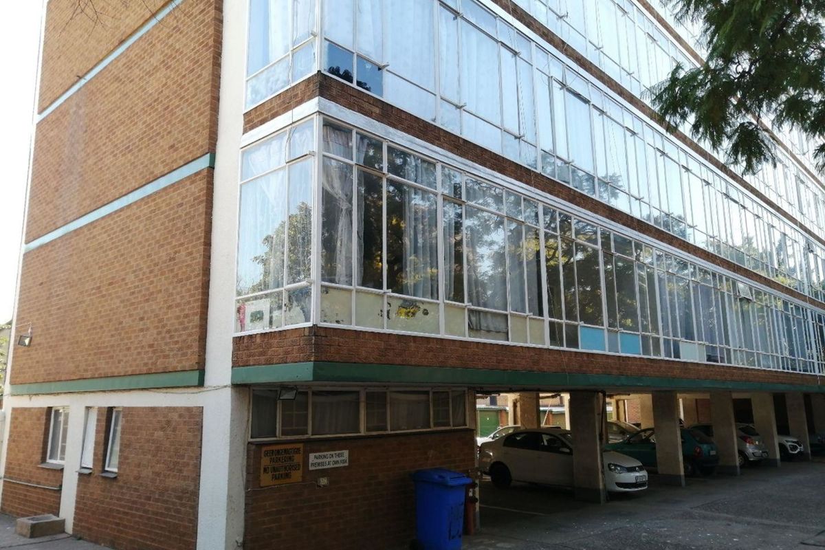 Exterior view of an apartment building featuring a brick facade, extensive glass window panels, and a ground-level covered parking area.
