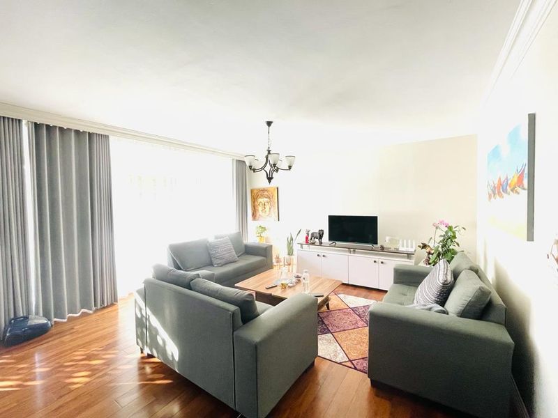 A bright and spacious living room featuring polished hardwood flooring, a central chandelier, and large windows with floor-to-ceiling curtains.