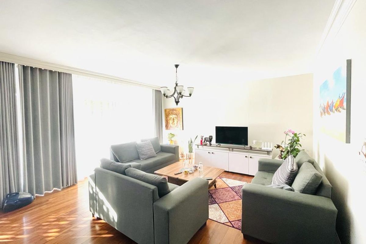 A bright and spacious living room featuring polished hardwood flooring, a central chandelier, and large windows with floor-to-ceiling curtains.