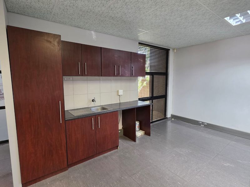 A functional office kitchenette featuring dark wood-finish cabinetry, a stainless steel sink, tiled backsplash, and durable floor tiles.