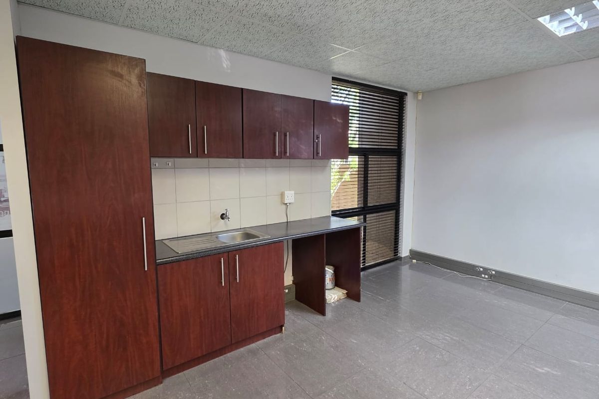 A functional office kitchenette featuring dark wood-finish cabinetry, a stainless steel sink, tiled backsplash, and durable floor tiles.