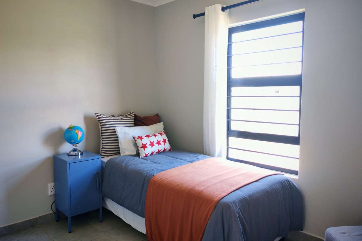 A functional bedroom featuring neutral wall tones, tiled flooring, and a large window with security bars providing natural light.