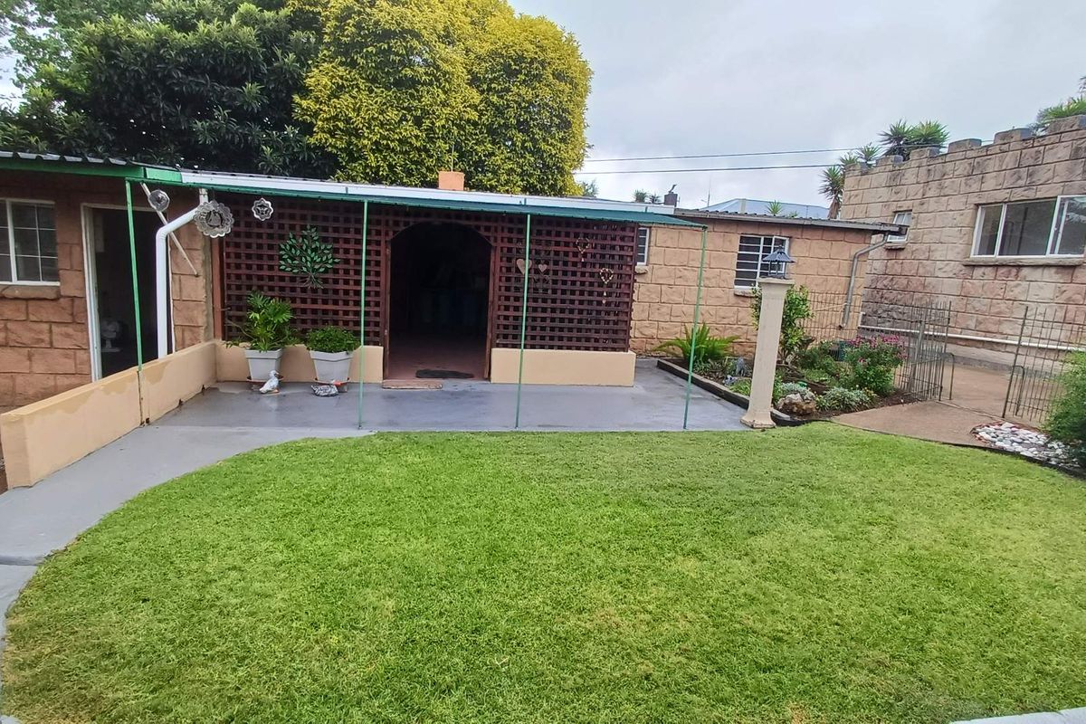 A rear garden view featuring a covered patio area with lattice screening, adjacent to a stone-clad house exterior and a well-maintained lawn.