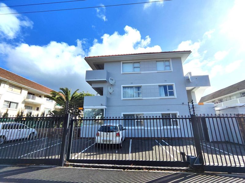 Front elevation of a multi-story apartment building featuring a paved parking area, secure metal gate, and electric fencing.