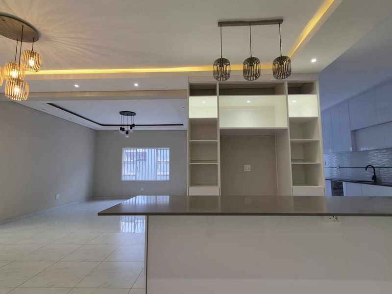 Modern open-plan living and kitchen area featuring a central island, built-in shelving, sleek cabinetry, and contemporary recessed and pendant lighting throughout.