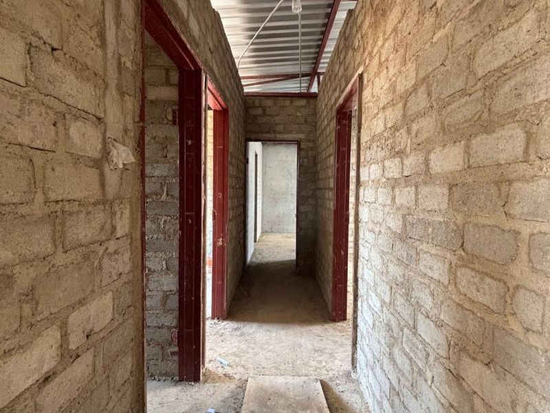 An unfinished hallway interior showing exposed brick walls, metal door frames, and an exposed roof structure, currently in the early stages of construction.
