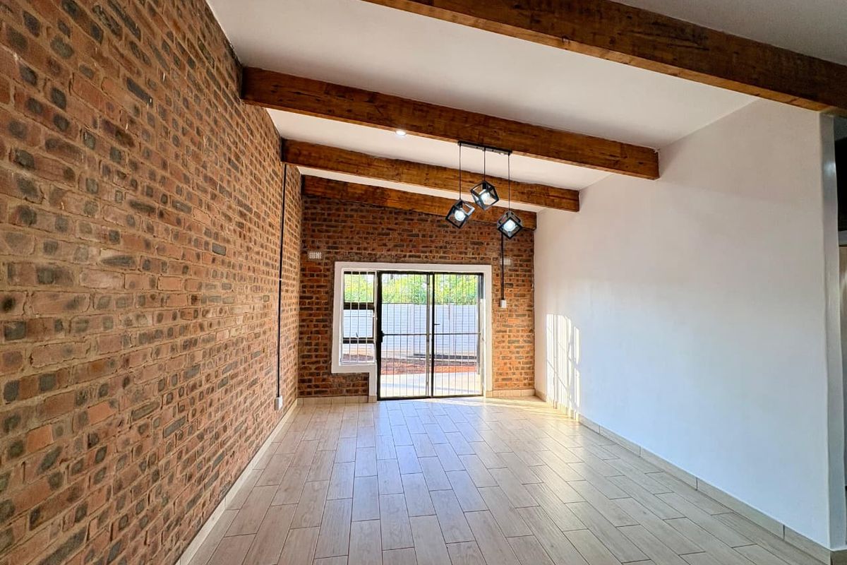 A bright living area featuring exposed brick walls, timber ceiling beams, tiled flooring, and a sliding door leading to the exterior.