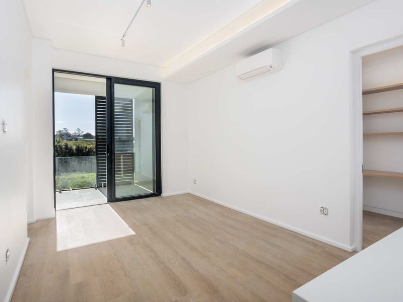 A bright, modern bedroom featuring light wood-look flooring, a split-system air conditioner, and a sliding glass door leading to a private balcony.