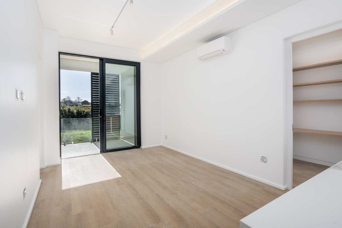 A bright, modern bedroom featuring light wood-look flooring, a split-system air conditioner, and a sliding glass door leading to a private balcony.
