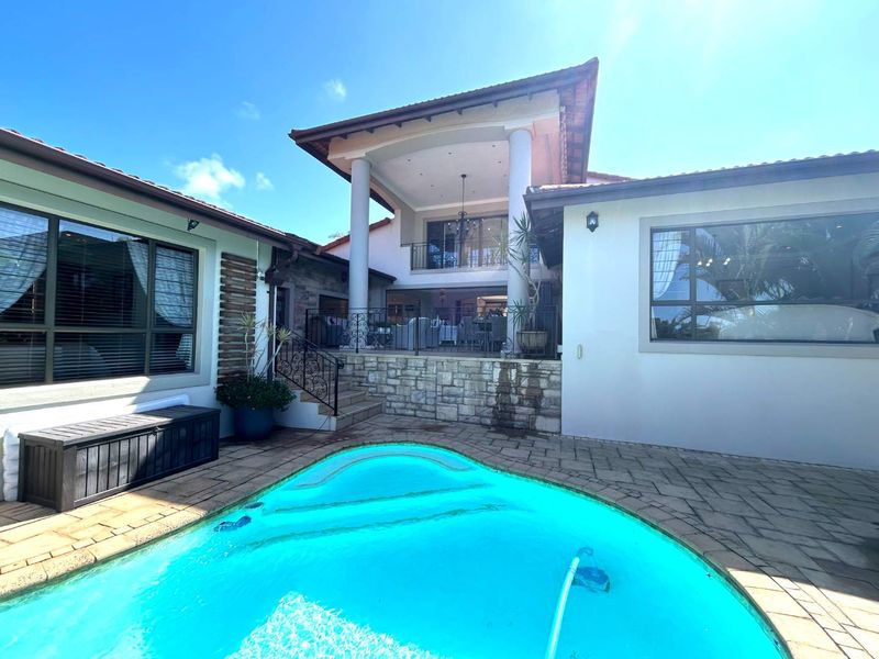 Rear exterior view featuring a swimming pool, paved deck, and a covered patio area with stone accents and wrought iron railings.