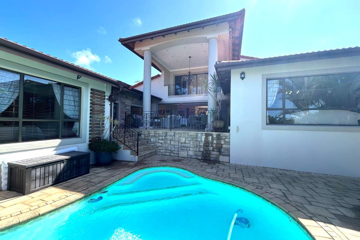 Rear exterior view featuring a swimming pool, paved deck, and a covered patio area with stone accents and wrought iron railings.