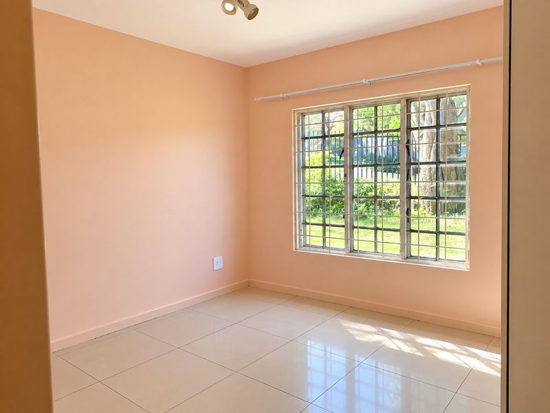 A bright, empty bedroom featuring neutral tiled flooring, peach-toned walls, and a window fitted with security bars and a curtain rail.