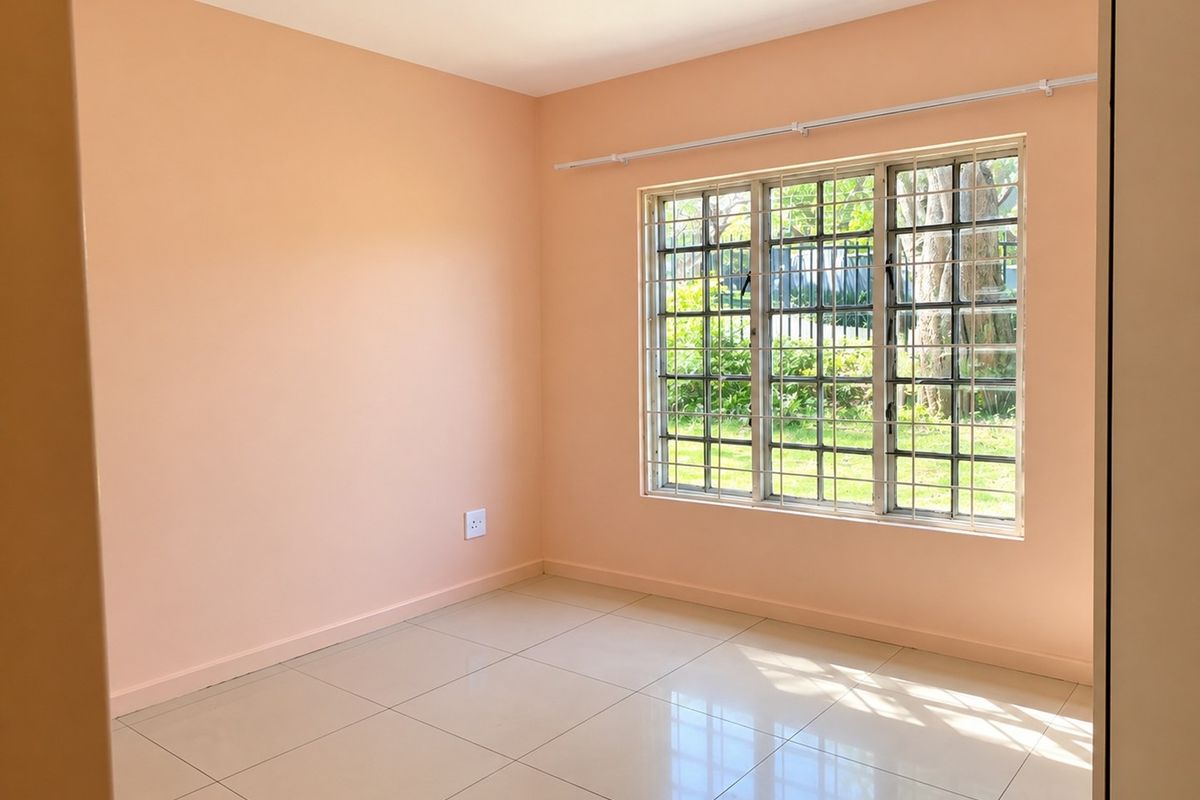A bright, empty bedroom featuring neutral tiled flooring, peach-toned walls, and a window fitted with security bars and a curtain rail.