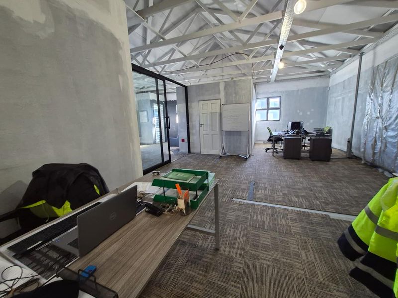 Open-plan office space featuring exposed roof trusses, carpet tile flooring, and a glass partition, currently undergoing renovation with unfinished wall surfaces.
