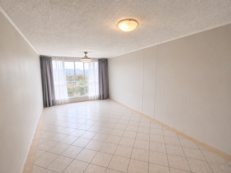 A spacious, empty living area featuring neutral tiled flooring, a ceiling fan, and a large window providing natural light and exterior views.