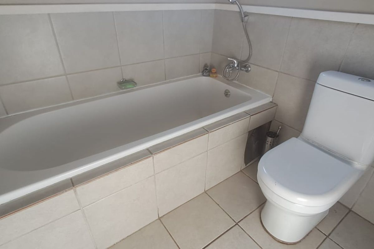 A functional bathroom featuring a built-in bathtub with shower mixer and a standard toilet, finished with neutral wall and floor tiling.