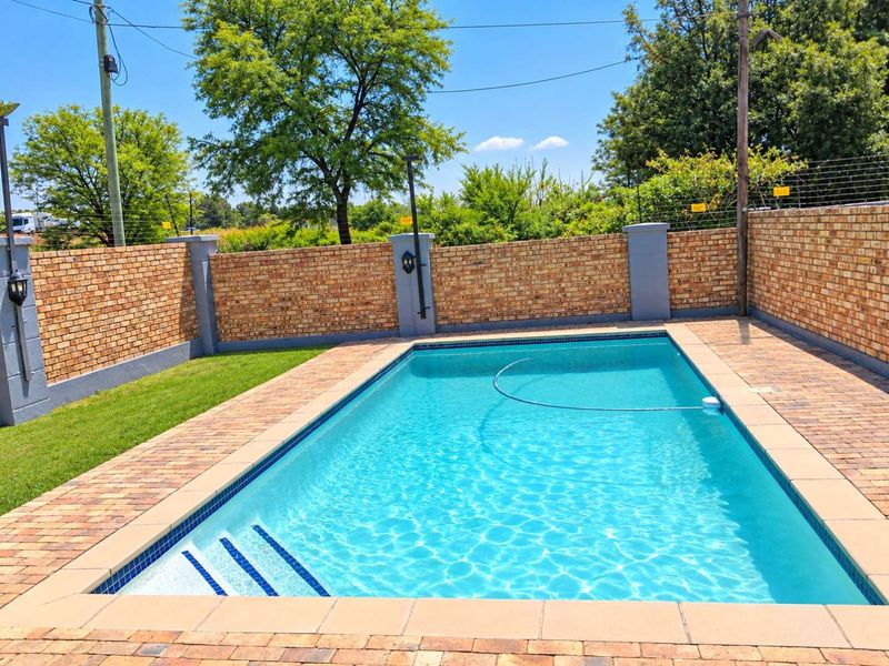 A well-maintained swimming pool area featuring a paved surround, manicured lawn, and secure brick boundary walls with electric fencing.