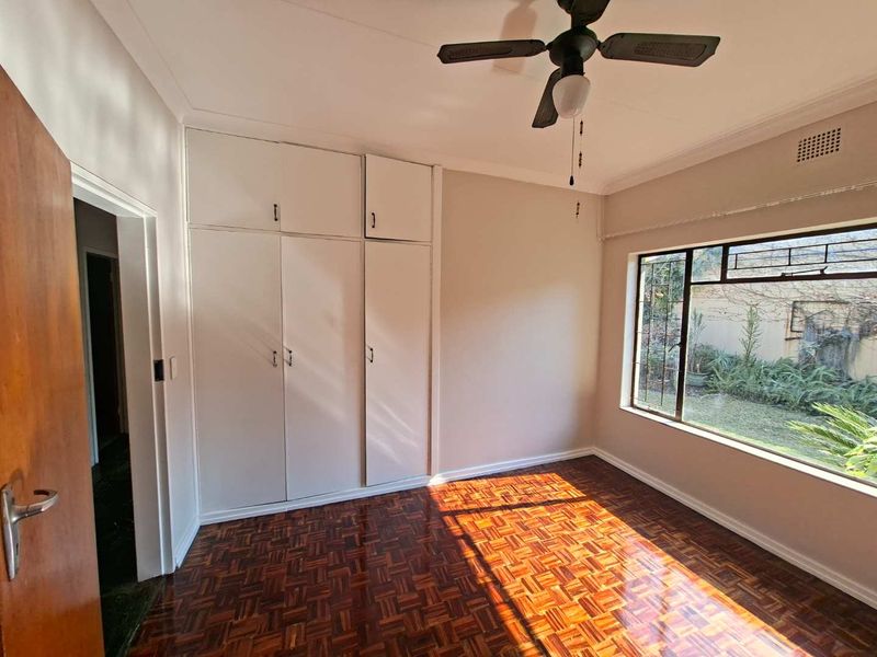 A bright, empty bedroom featuring classic parquet flooring, built-in floor-to-ceiling cupboards, a ceiling fan, and a large window overlooking the garden.
