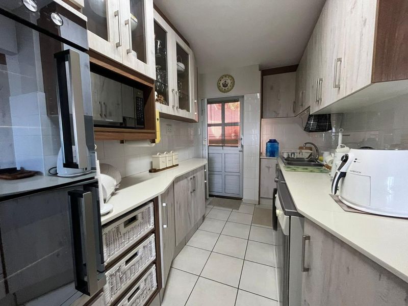 A functional galley-style kitchen featuring light wood-grain cabinetry, laminate countertops, tiled flooring, and built-in appliances with direct access to an exterior door.