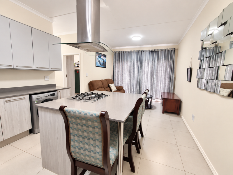 Modern open-plan living area featuring a central kitchen island with a gas hob, stainless steel extractor, and sleek cabinetry, flowing into a bright living space.