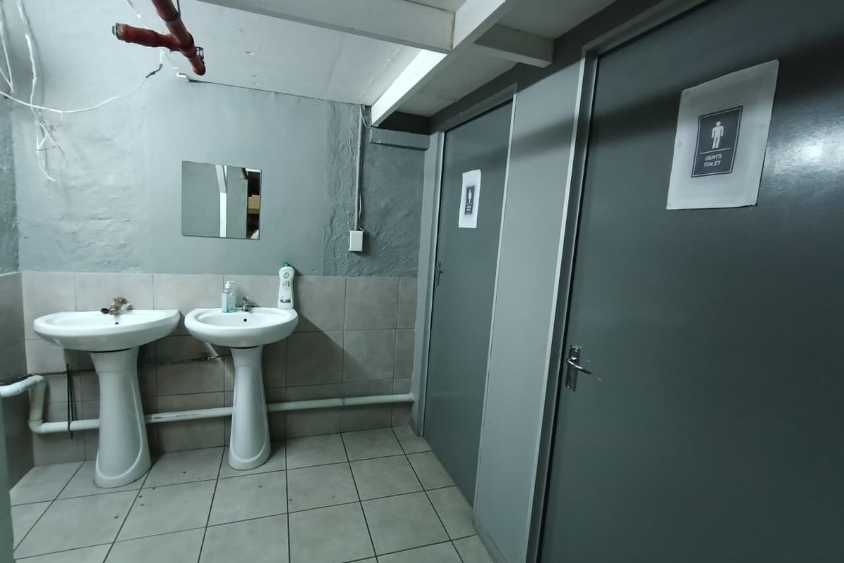 Industrial style bathroom facility featuring dual pedestal wash basins, tiled flooring, and access to separate toilet cubicles with exposed overhead fire safety infrastructure.