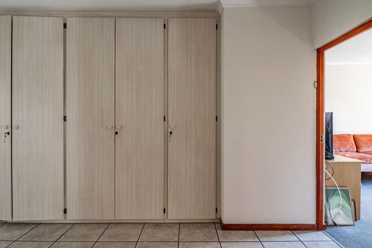 A bedroom featuring light wood-grain built-in cupboards and tiled flooring, with a view into an adjacent room through a timber-framed doorway.