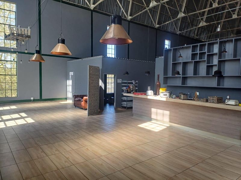 Spacious open-plan industrial-style commercial space featuring high ceilings with exposed trusses, large windows, tiled flooring, and a built-in bar area with shelving.