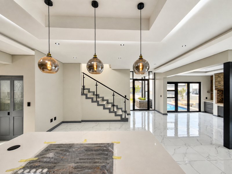 Spacious open-plan living area featuring modern pendant lighting, a sleek staircase with glass balustrade, polished marble-look flooring, and seamless access to the outdoor pool area.