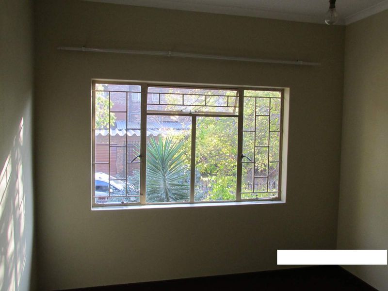 An empty bedroom featuring a large window with security bars and a curtain rail, providing natural light and a view of the exterior.