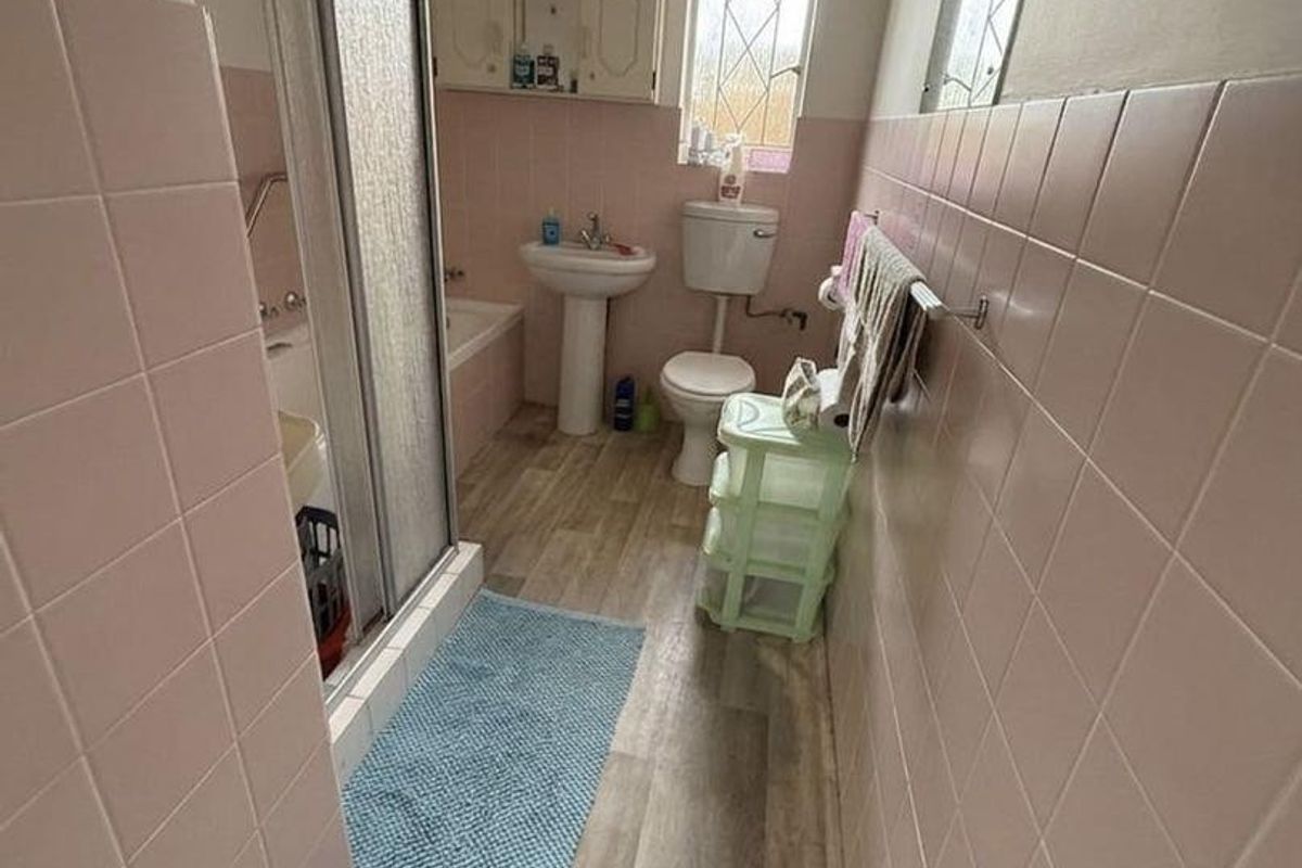 A dated bathroom featuring a shower enclosure, pedestal basin, and toilet, finished with pink wall tiles and wood-look flooring.