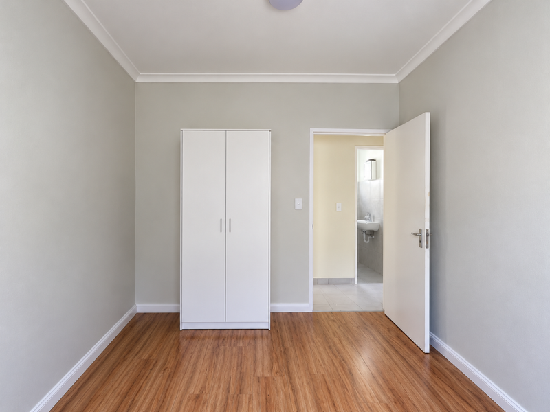 A clean, empty bedroom featuring laminate flooring, neutral walls, coving, and a built-in wardrobe, with an adjacent bathroom visible through the open doorway.
