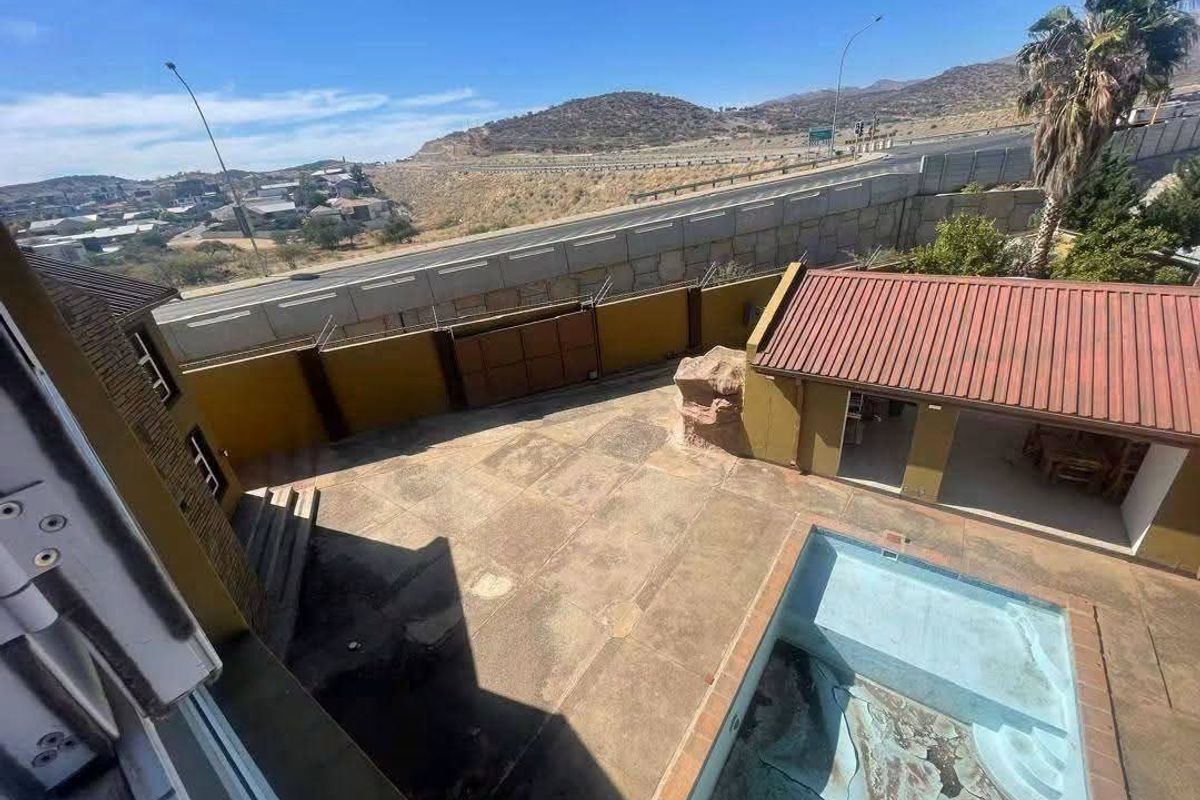 Aerial view of a backyard featuring a swimming pool, paved patio area, and a detached outbuilding, enclosed by a high boundary wall with security spikes.