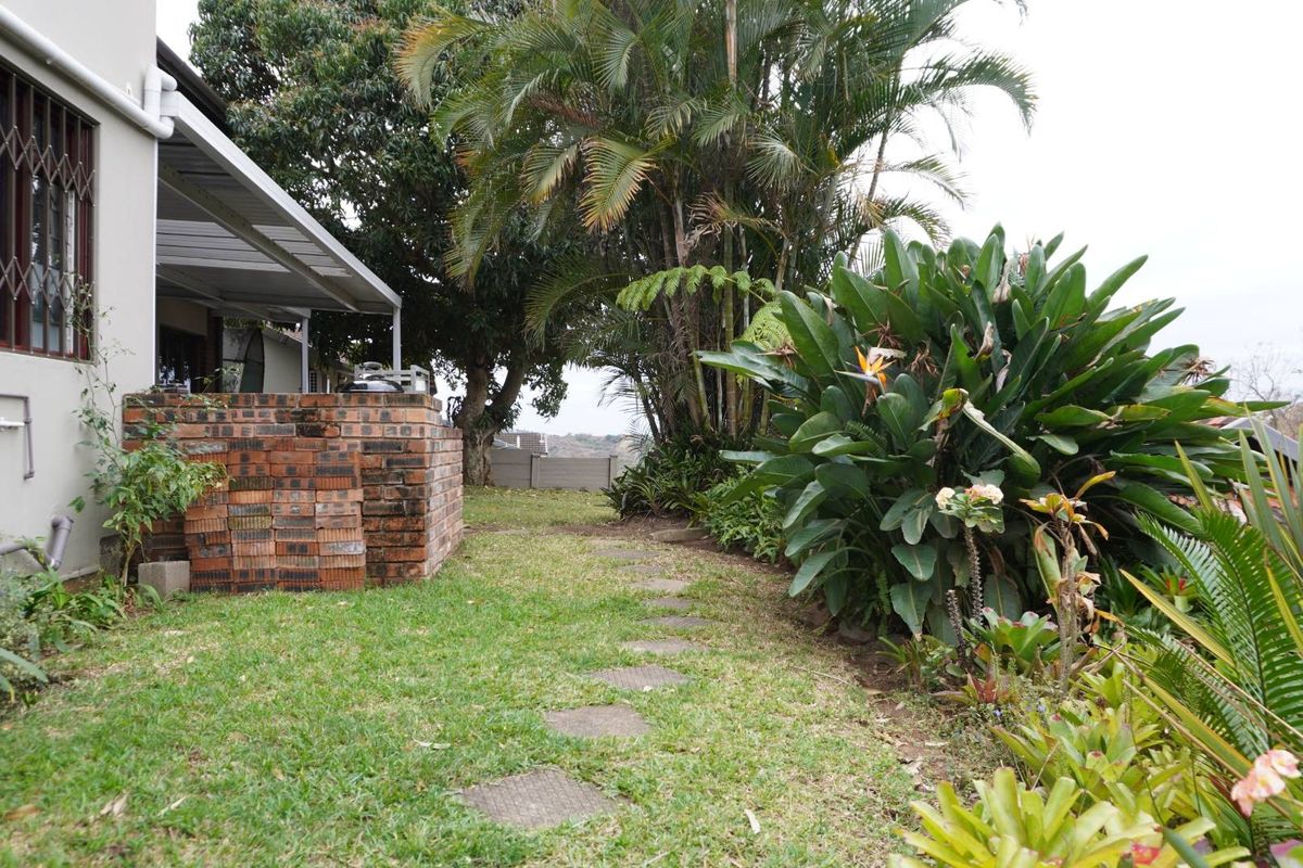 Rear garden area featuring a brick built-in braai, paved stepping stones, and established garden beds with mature trees and shrubs.