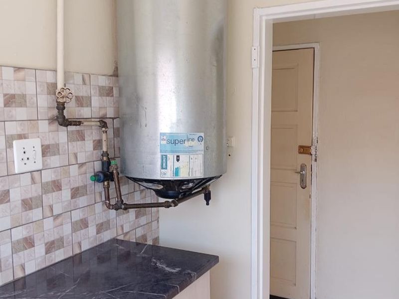 Utility area featuring a wall-mounted electric water heater, dark laminate countertop, base cabinetry, and tiled splashback, leading to an adjacent room.