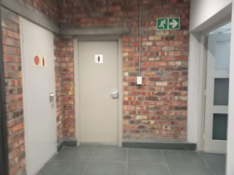 A functional office hallway featuring exposed brick walls, tiled flooring, and clear emergency exit signage leading to restroom facilities.