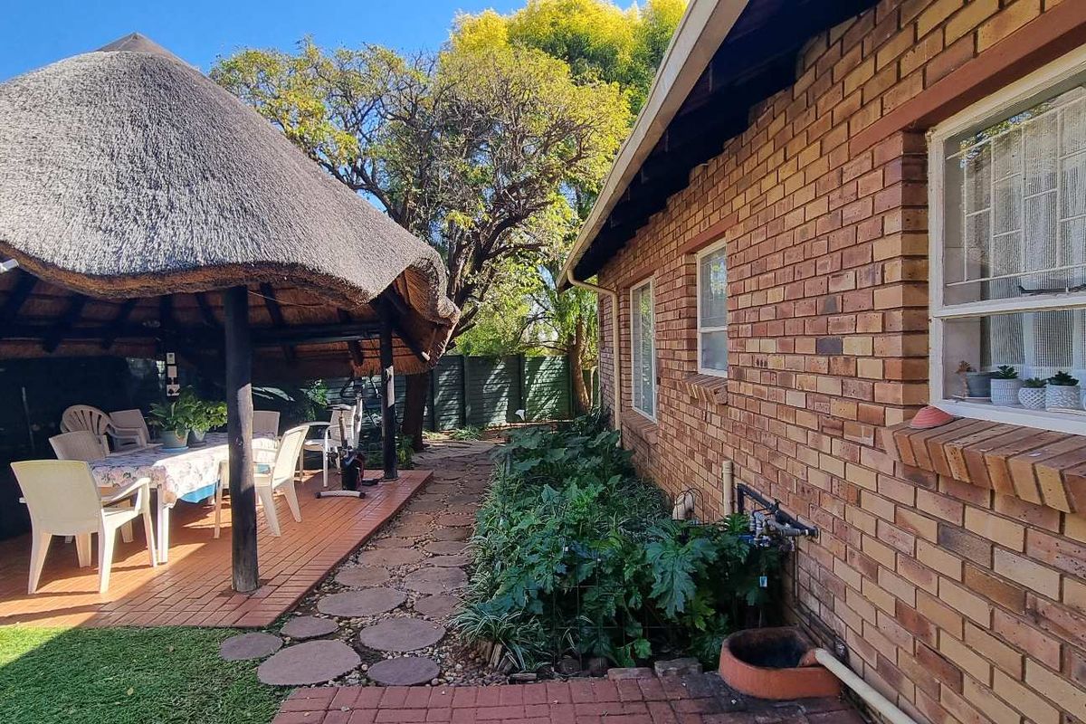 A rear garden area featuring a traditional thatched lapa, brick-paved patio, and established garden beds alongside the main house structure.