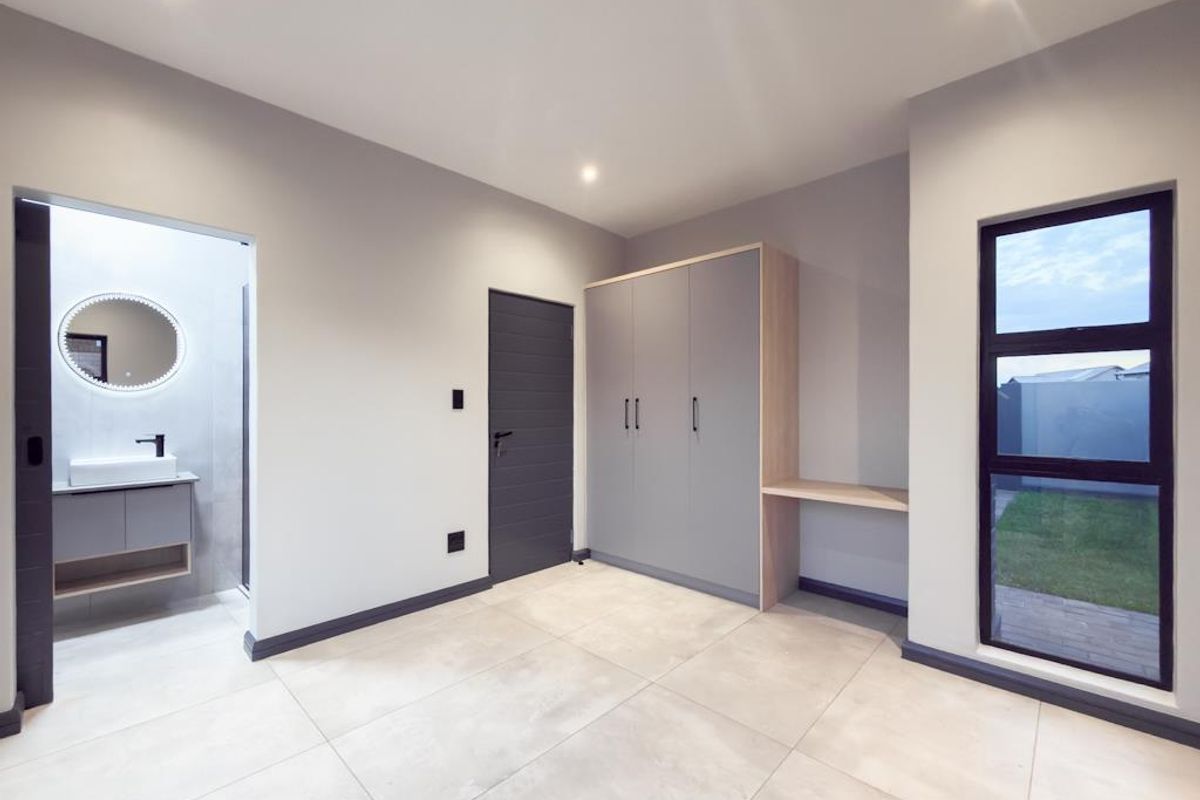 A modern, well-lit bedroom featuring neutral floor tiles, built-in cabinetry with a desk area, and an en-suite bathroom with a contemporary vanity.