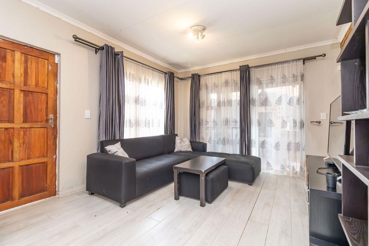 A living room featuring light-toned laminate flooring, a solid wood entrance door, and large windows with curtain tracks.