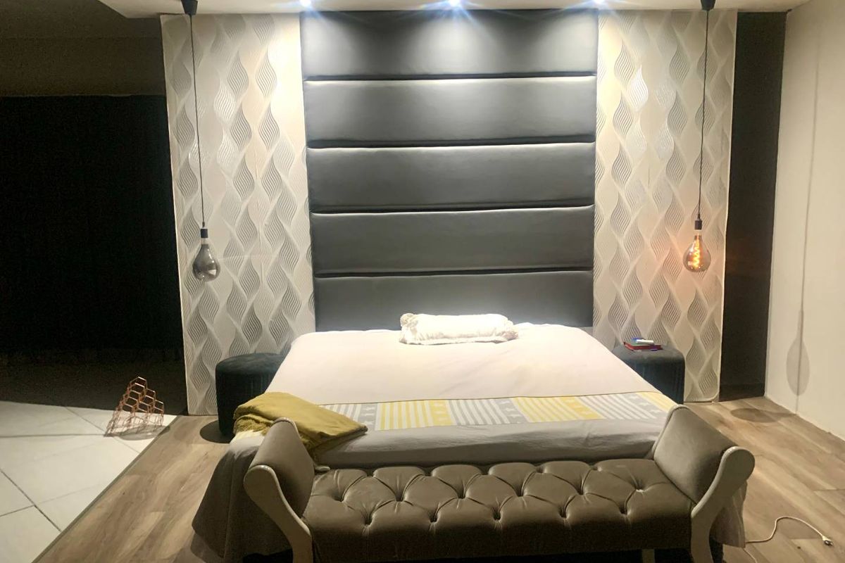 A modern bedroom featuring a large upholstered headboard, recessed ceiling lighting, and stylish pendant lights, complemented by wood-look laminate flooring.